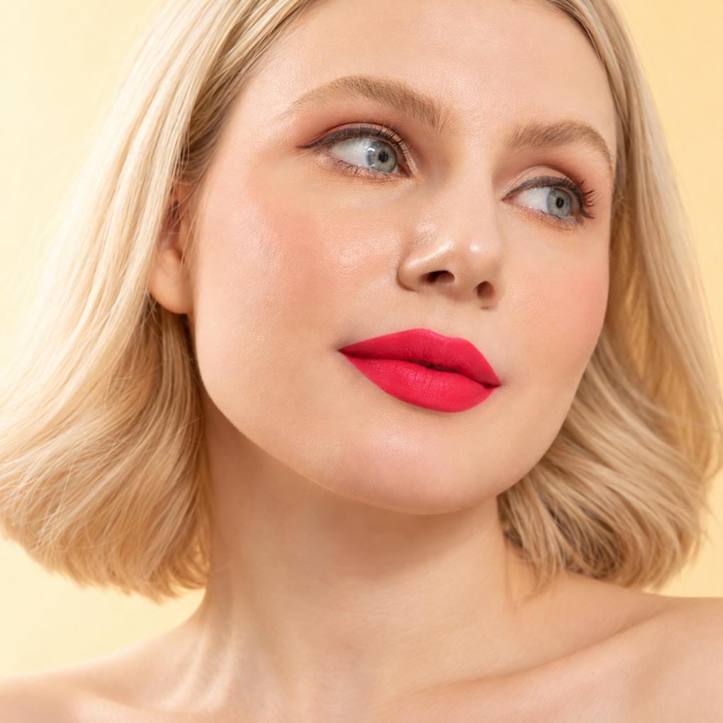 Model wearing Oplenza Signature Longwear Matte Lipstick in 11 Garnet, with bright pink-red lipstick, looking upward against a soft warm beige background.